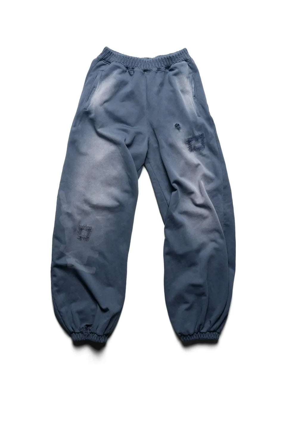 Racer WorldwideのBat Wing Sweatpantsのフロントビュー。外側が湾曲した立体的なバットウィングシルエットが特徴のウォッシュドブルースウェットパンツ。太腿や膝付近にダメージとリペアパッチワークが施されている。