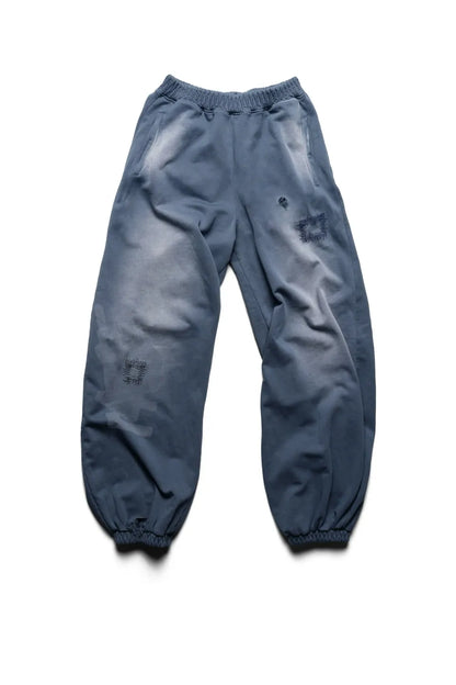 Racer WorldwideのBat Wing Sweatpantsのフロントビュー。外側が湾曲した立体的なバットウィングシルエットが特徴のウォッシュドブルースウェットパンツ。太腿や膝付近にダメージとリペアパッチワークが施されている。